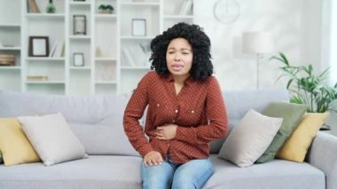 Genç Afro-Amerikan kadın karın ağrısından muzdarip evdeki kanepede oturuyor. Üzgün siyah kadın ellerini karnında tutuyor. Zehirlenme, kabızlık, spazm ya da regl olmuş.