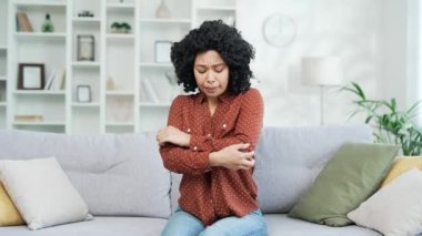Evdeki oturma odasındaki kanepede otururken dirsek ağrısından muzdarip Afro-Amerikalı bir kadın. Üzgün siyah kadın bir eklem masajı yapıyor, elini ovuyor, eklemlerinde rahatsızlık hissediyor.