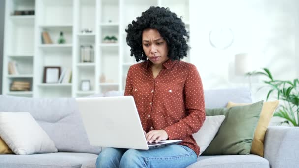 失望的年轻非洲裔美国女性坐在家里的沙发上 在笔记本电脑上阅读坏消息 伤心的黑人女人因不愉快的通知而烦恼 因信息而震惊 因信息而沮丧 — 图库视频影像