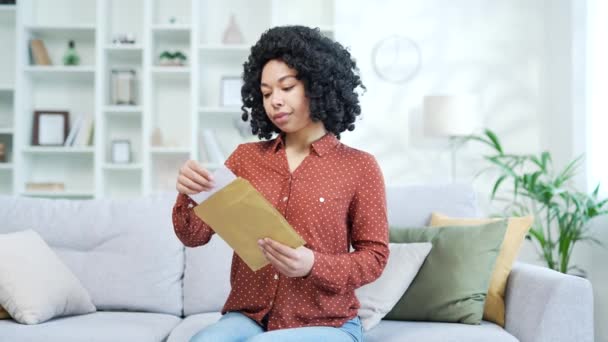快乐而兴奋的年轻的非洲裔美国女性 坐在家里的沙发上 阅读着有好消息的信 快乐的微笑的黑人女人满足于一个愉快的通知 庆祝成功 — 图库视频影像