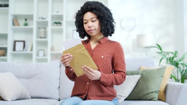 若いアフリカ系アメリカ人女性が 自宅でソファーに座っているという悪いニュースを読んでいるのを失望させた 悲しい黒人女性は不快な通知に落ち込んでおり 文書内の情報にショックを受けています — ストック動画