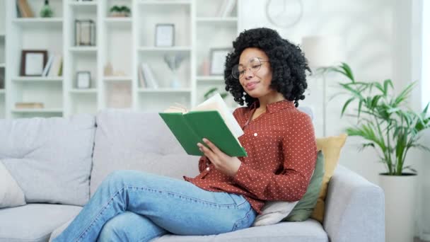 年轻快乐的非洲裔美国女人坐在客厅的沙发上看书 一个戴着眼镜的微笑的黑人女人在闲暇时阅读她最喜欢的文学作品 学生们正在用课本学习 — 图库视频影像