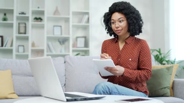 年轻的非洲裔美国女学生自由职业者坐在家里的办公室里听在线电子学习 视频课程 参加远程学习研讨会的黑人妇女用笔记本电脑在笔记本上写字 — 图库视频影像