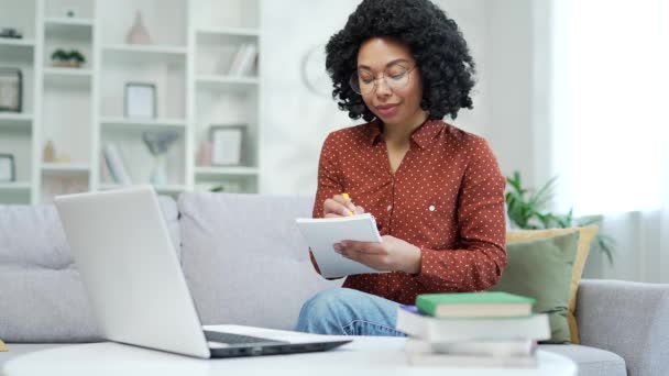 年轻的非洲裔美国女学生自由职业者坐在家里的办公室里 用笔记本电脑听在线电子学习 远程学习中的黑人妇女在笔记本上写字 与家庭教师交流 — 图库视频影像