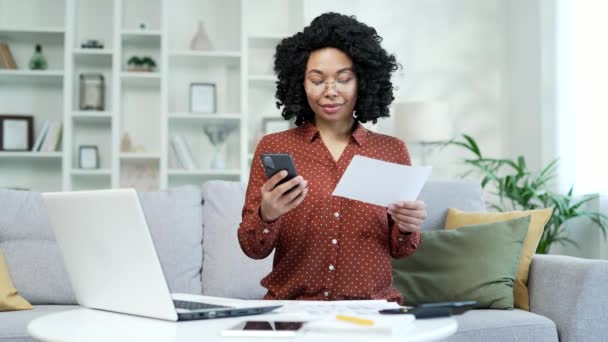 年轻的非洲裔美国女人坐在家里用智能手机上的网站或应用程序处理家庭财务 黑人妇女通过在电话上输入数据 持有公用事业费账单 并在网上付款 — 图库视频影像