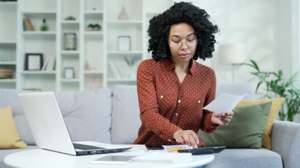 年轻的非洲裔美国女人在家里的客厅里坐在沙发上处理家庭财政问题 黑人妇女在计算器上计算 在笔记本上输入数据 在笔记本电脑上输入数据 — 图库视频影像