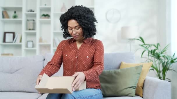 一位兴奋的非洲裔美国女性顾客打开了一个包裹回家 快乐的黑人妇女买主坐在沙发上打开包装购买 电子商务的满意客户 速递服务概念 — 图库视频影像