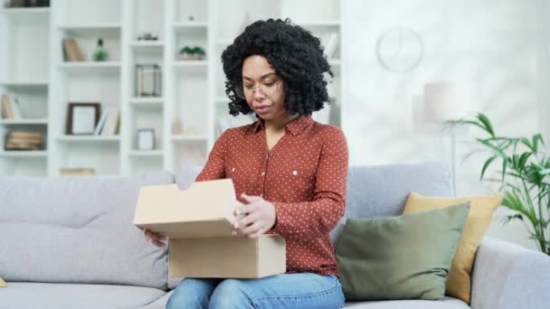 不幸的非洲裔美国女人在家里客厅的沙发上收到了一个装了错误产品的包裹 黑人妇女对在网上商店购物感到不满 服务问题或受损物品 — 图库视频影像