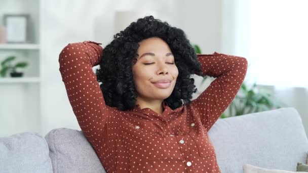 アフリカ系アメリカ人の若い女性が目を閉じてリラックスし リビングルームに座っている間に頭の後ろに手を置いた 自宅で仕事をした後に休む落ち着いた幸せな黒人女性の肖像画 — ストック動画