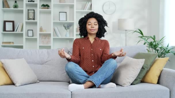 年轻的非洲裔美国女性坐在客厅的沙发上 闭着眼睛沉思着 一个沉着快乐的黑人女人做瑜伽 深呼吸 在舒适的沙发上放松 — 图库视频影像