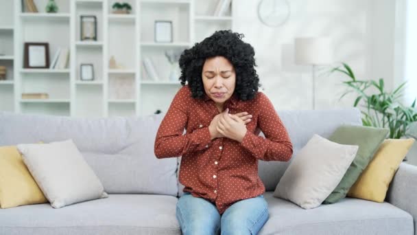 胸の張力に苦しむ病気の若いアフリカ系アメリカ人女性 自宅のリビングルームでソファーに座っている心臓発作 彼女の手で胸を持っている黒人女性は 緊急の医療を必要とします — ストック動画