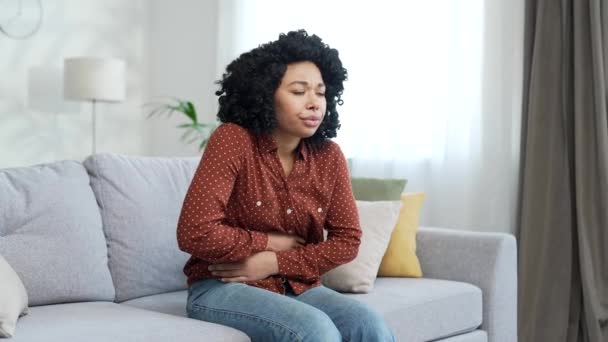 腹痛に苦しむアフリカ系アメリカ人女性が 自宅の部屋でソファーに座っている 悲しい黒人女性は彼女の胃に手を置いています 彼女は中毒 スパムまたは月経を持っています — ストック動画
