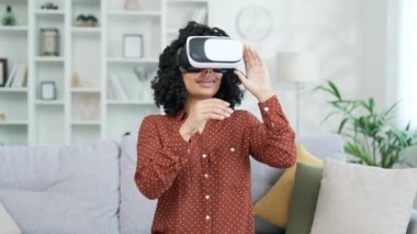 Genç başarılı Afro-Amerikan kadın sanal gerçeklik simülatöründe VR gözlük kullanarak çalışıyor. Evdeki koltukta oturuyor. Kadın bilgisayar programlarını kontrol etmek için jestler kullanır ve sanal sayfaları çevirir