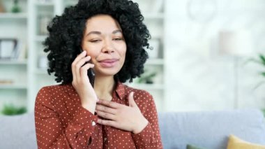 Kapatın. Evdeki oturma odasında kanepede otururken akıllı telefondan konuşan mutlu Afro-Amerikan kadın. Gülümseyen siyah kadın bir meslektaşıyla konuşuyor ya da bir arkadaşıyla iletişim kuruyor.