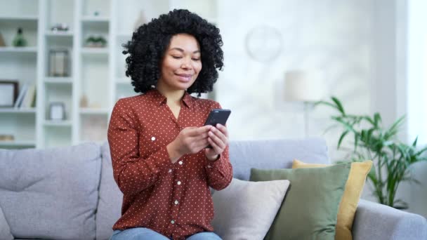 年轻的非洲裔美国女性坐在客厅的沙发上 使用智能手机 微笑的黑人女人浏览社交网络 在网上商店购物 写作或阅读信息 — 图库视频影像