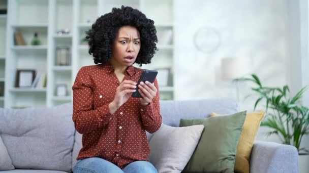スマートフォンで悪いニュースを読んでいる若いアフリカ系アメリカ人女性が 自宅のリビングルームでソファーに座っていることに失望した 悲しいショックを受けた黒人女性は不快な通知に動揺し メッセージによって落ち込んだ — ストック動画