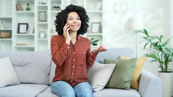 快乐的年轻非洲裔美国女性坐在客厅的沙发上 一边用智能手机聊天 微笑的黑人女人与同事交谈或与朋友男朋友交流 — 图库视频影像