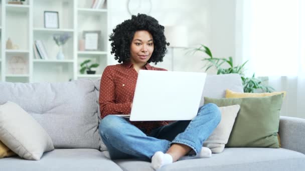 年轻的非洲裔美国女自由职业者坐在家里的沙发上打字 黑人女学生在电脑上工作 与朋友在网上聊天 写电子邮件或浏览社交媒体 — 图库视频影像