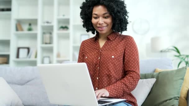年轻的非洲裔美国女自由职业者坐在总公司的沙发上 在笔记本电脑上打字 黑人女企业家在电脑上的一个项目上工作 与客户在网上聊天 写电子邮件 — 图库视频影像