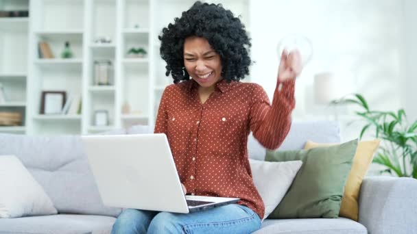 快乐的年轻非洲裔美国女性通过在家里客厅的笔记本电脑上阅读好消息来庆祝成功 兴奋地微笑着的黑人女人 对坐在沙发上的愉快信息感到满意 — 图库视频影像