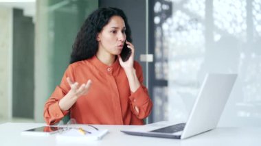 Modern ofisteki çalışma masasında otururken kendine güvenen genç bir bayan çalışanın akıllı telefonuyla konuşması. Ciddi esmer bir kadın ortağıyla iş konuşur ya da müşterisiyle konuşur.