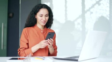 Genç bayan çalışan modern ofisteki iş yerinde otururken akıllı telefon kullanıyor. Gülümseyen kıvırcık esmer kadın mesaj yazıyor, internette sohbet ediyor, kaydırıyor, sosyal medyada geziniyor ya da e-postaları kontrol ediyor.