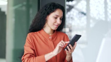 Genç bayan çalışanlar modern ofisteki iş yerinde otururken akıllı telefon kullanıyorlar. Gülümseyen kıvırcık esmer kadın sosyal medyada geziniyor, online alışveriş yapıyor, mesaj yazıyor ya da sohbet ediyor. 