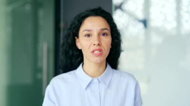 Webcam görüntüsü. Kapatın. Genç bayan çalışan, iş yerinde kameraya bakarak bir video görüşmesinde konuşuyor. Kendine güvenen koç uzaktan konuşur, çevrimiçi bir konferansta bir konuşma yapar.