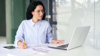 Ciddi genç kadın muhasebeci, iş yerinde masa başında otururken dizüstü bilgisayar kullanarak not alıyor. Düşünceli muhasebeci ya da finansör hesaplar oluşturur, modern ofiste görevleri düşünür.