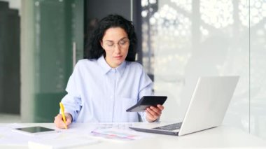 Meşgul genç kadın muhasebeci dizüstü bilgisayar ve hesap makinesi kullanarak hesaplar yapar, iş yerinde otururken notlar alır. Modern ofiste çalışan kendine güvenen muhasebeci ya da gözlüklü yatırımcı
