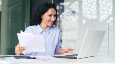 Mutlu genç kadın finansör, bilgisayardaki ekrana bakarak ve ofisteki bir iş yerinde duran belgeleri tutarak başarıyı kutluyor. Girişimci olumlu, iyi mali sonuçlardan memnun