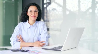 Modern bir ofiste işyerinde oturan, gözlüklü, gülümseyen genç bir kadın çalışanın portresi. Tişörtlü mutlu kıvırcık esmer kadın kameraya bakıyor. Başarılı bir menajerin vesikalığı