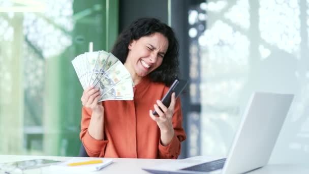 快乐而兴奋的年轻女性员工一边挥动着钞票 一边看着坐在现代办公室工作场所的智能手机屏幕 微笑的女人手里拿着电话和钞票 看着相机 — 图库视频影像