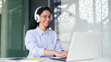 Modern ofisteki çalışma masasında otururken kulaklıkla dizüstü bilgisayarda yazan genç ve mutlu bayan çalışan. Gülümseyen kıvırcık saçlı esmer kadın en sevdiği müziği dinlerken bilgisayarda çalışıyor.