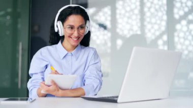 Kulaklıklı kadın çalışan, ofiste otururken internetten e-öğrenme videosu dinliyor. Uzaktan öğrenme seminerindeki bir kadın dizüstü bilgisayarla koçla iletişim kurar.