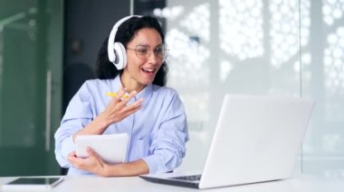 Kulaklıklı kadın çalışan, ofiste otururken internetten e-öğrenme videosu dinliyor. Uzaktan öğrenme seminerindeki bir kadın dizüstü bilgisayarla koçla iletişim kurar.