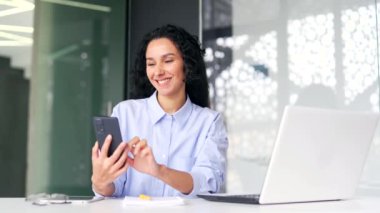 Genç bayan çalışan modern ofisteki iş yerinde otururken akıllı telefon kullanıyor. Gülümseyen kıvırcık esmer kadın mesaj yazıyor, sohbet ediyor, kayıyor, sosyal medyada geziniyor ya da internetten alışveriş yapıyor.