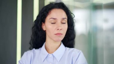 Kapatın. Modern bir ofiste duran kendine güvenen genç bir kadın çalışanın portresi. Mavi gömlekli esmer, ciddi bir kıvırcık kadın kameraya bakıyor. Başarılı bir menajerin vesikalığı