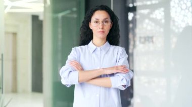Modern bir ofiste kolları çapraz duran mavi gömlekli, kendine güvenen genç bir kadın çalışanın portresi. Gözlüklü esmer kadın kameraya bakıyor. Başarılı bir menajerin vesikalığı