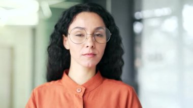 Kapatın. Modern bir ofiste duran kendine güvenen genç bir kadın çalışanın portresi. Gözlüklü ciddi kıvırcık esmer kadın kameraya bakıyor. Başarılı bir menajerin veya ajanın vesikalığı