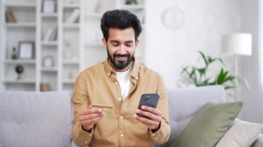 Mutlu genç sakallı adam online alışveriş yapıyor. Evdeki kanepede oturan akıllı telefondan kredi kartı numarasını yazıyor. Tişörtlü, gülümseyen yakışıklı bir erkek, ürün tanıtımından memnun bir şekilde satın alıyor.