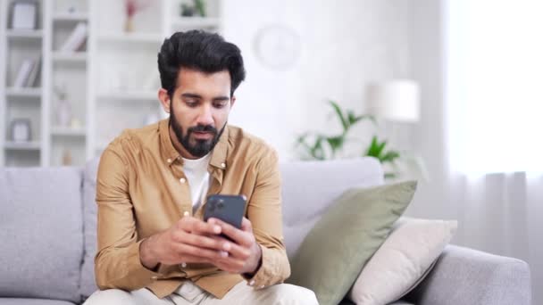Junger Bärtiger Mann Sitzt Mit Smartphone Auf Sofa Heimischen Wohnzimmer — Stockvideo