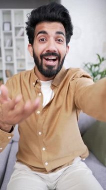 POV Webcam görüntüsü. Dikey video. Genç yakışıklı bir adam evdeki kanepede otururken akıllı telefon kullanarak bir video görüşmesinde konuşuyor. Gülümseyen pozitif sakallı bir erkek bir arkadaşıyla veya meslektaşıyla internette sohbet eder.
