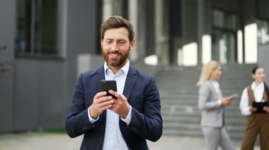Modern ofis binasının yakınında cep telefonu kullanarak yürüyen yakışıklı sakallı bir iş adamı. Açık havada, resmi takım elbiseli yakışıklı erkek yönetici dışarıda mesajlaşarak sohbet ediyor. Gülümse.