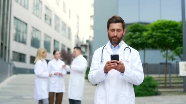 在诊所或医院办公室附近散步时 使用手机的成年男医生 男医生去浏览聊天打字短信阅读智能手机 — 图库视频影像