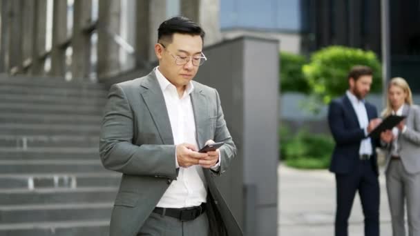 自信的亚洲商人使用手机在市中心大楼附近的城市街道上行走 商业成熟的男性创业者执行经理员工或穿着西服的老板外出 — 图库视频影像