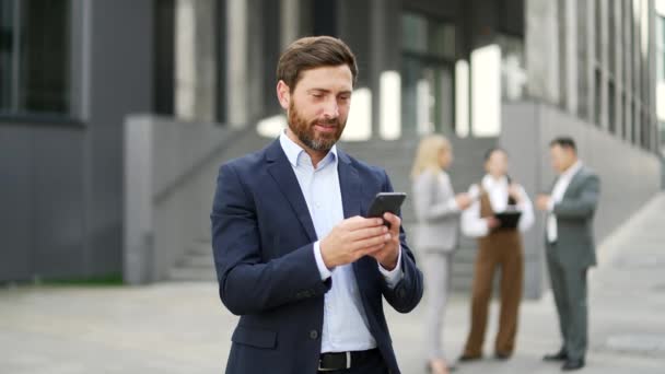 携帯電話を使用して近代的なオフィスの建物の近くに立っているハンサムなひげ付きビジネスマン屋外のスマイルの外でテキストをチャットする正式なスーツのブラウジングの魅力的な成熟した男性マネージャー — ストック動画
