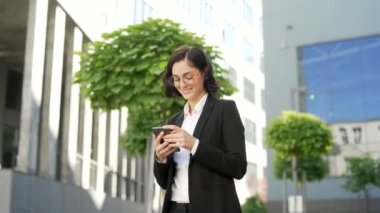Resmi takım elbiseli gülümseyen genç işadamı, cadde boyunca bir ofis binasının yakınında yürürken akıllı telefon kullanıyor. Kadın yönetici mesajlaşıyor, e- postaları kontrol ediyor, çevrimiçi sohbet ediyor veya mesajları okuyor