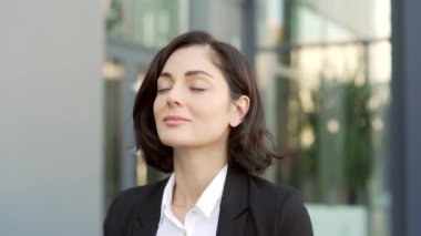 Kapatın. Mutlu genç iş kadını ofis binasının yanındaki sokakta gözleri kapalı dinleniyor. Resmi takım elbiseli neşeli esmer kadın derin derin nefes alır ve kameraya bakmaktan zevk alır.