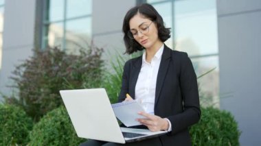 Resmi takım elbiseli bir kız öğrenci online webinar, sınıf web yayınları izliyor, dizüstü bilgisayara bakıyor, e-öğreniyor, notlar alıyor, video görüşmeleri yapıyor, kampüste bankta oturuyor, uzaktan öğretmen koçuyla konuşuyor.